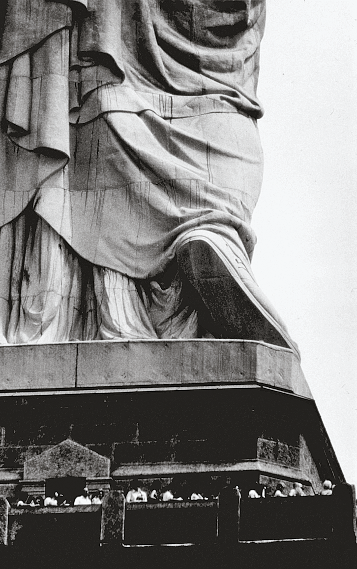 Photograph depicting pedestal of large sculpture with people standing around its base.