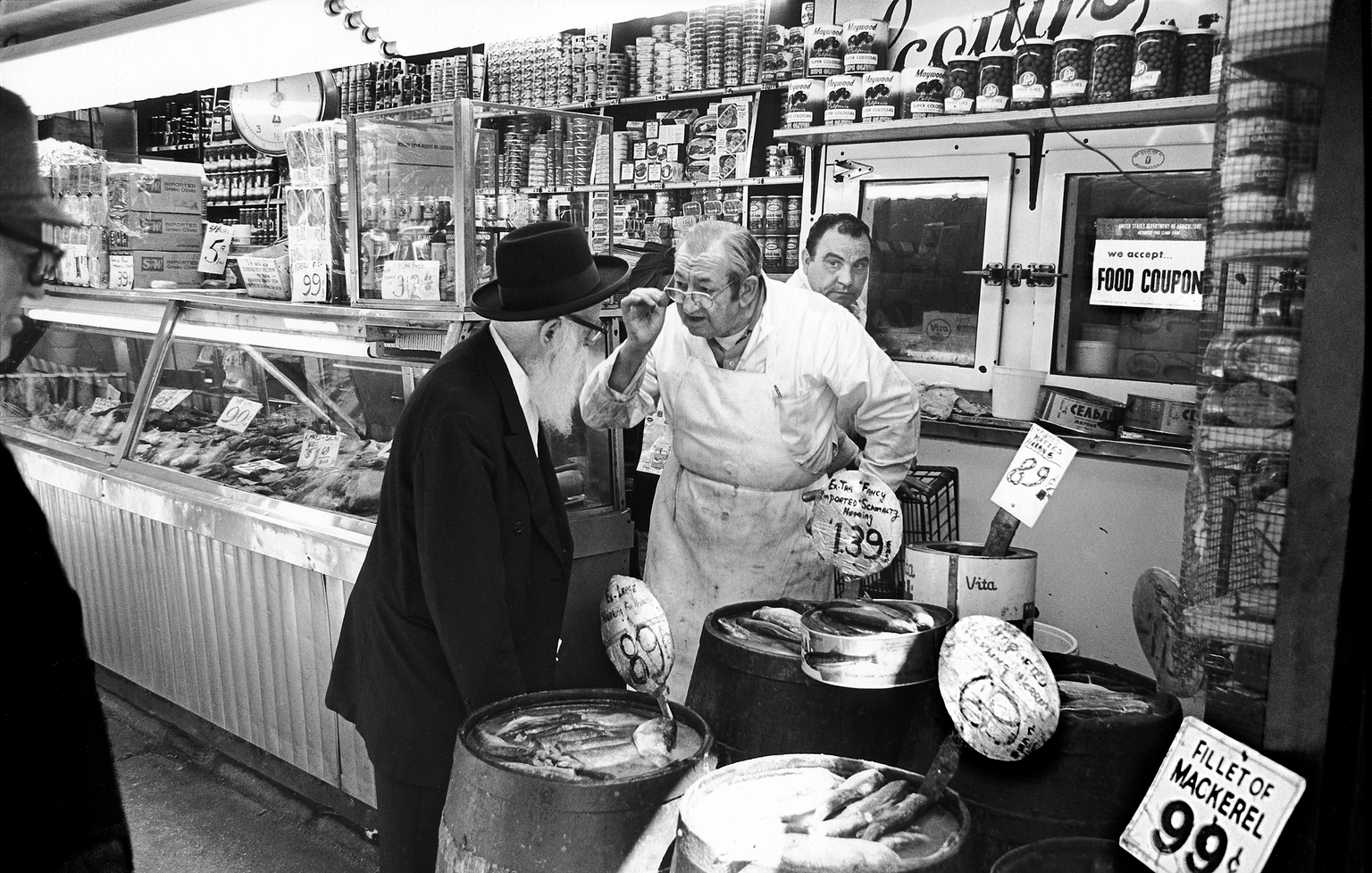 Photograph of interior of store with fish behind a glass counter and foodstuffs on shelves, with a man in a butcher's apron in center talking to a bearded man in a suit next to several barrels of fish and another man behind them.