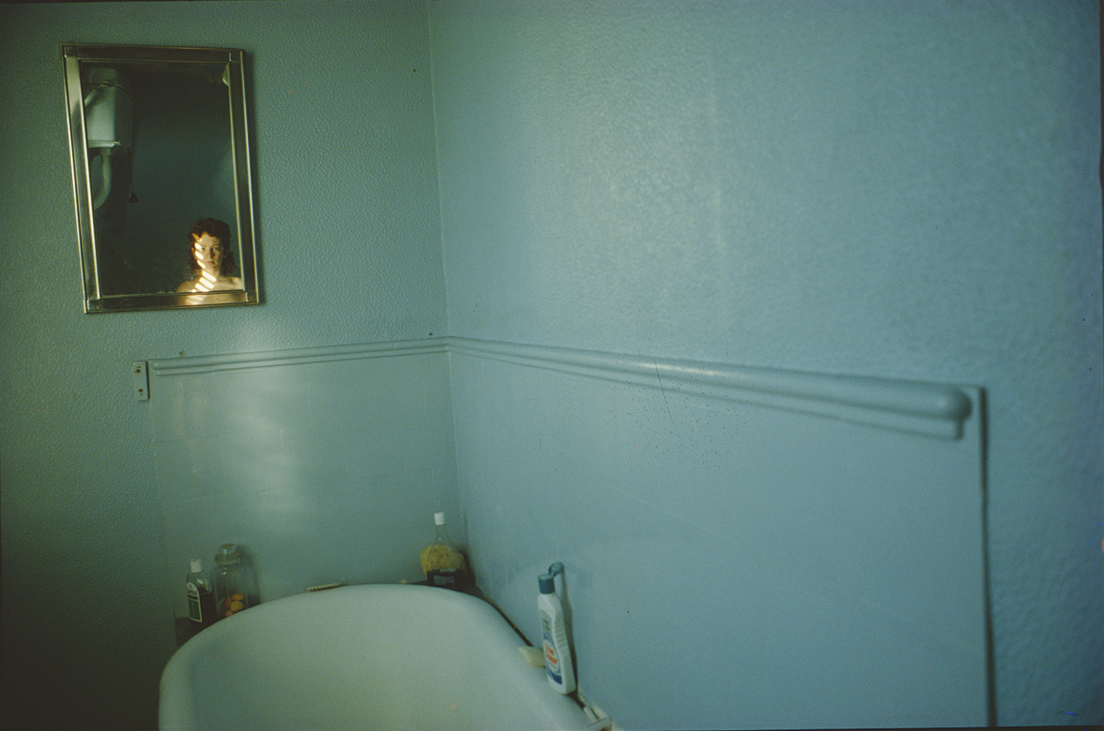 Photograph of bathroom wall with bathtub and small mirror above that reflects a face. 