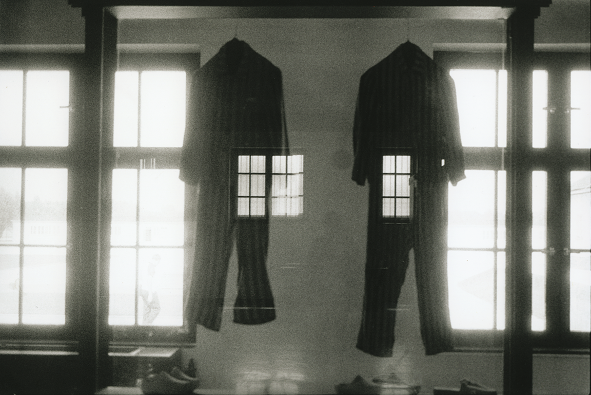 Photograph depicting two jumpsuits hanging between two large windows superimposed over an image of a human figure standing with their back turned. Photograph depicting two jumpsuits hanging between two large windows superimposed over an image of a human figure standing with their back turned.