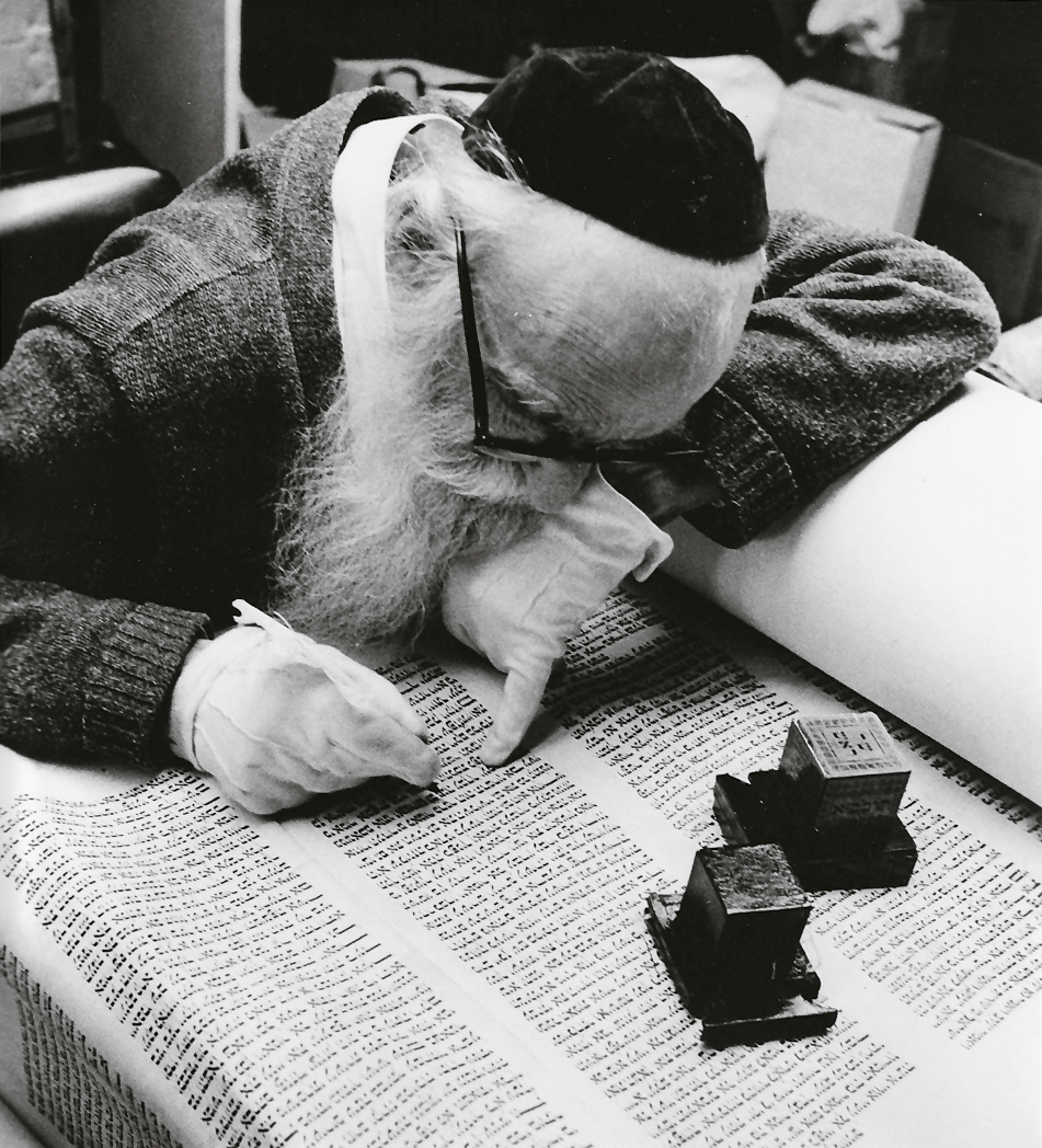 Photograph depicting man in kippah, gloves, and glasses and holding a pen examining a Torah scroll upon which sits two tefillin boxes.  