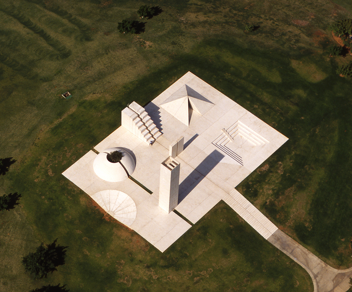 Concrete sculpture featuring a pyramid, water channel, tower with wind flutes, and a dome with olive tree in center set in a field. 