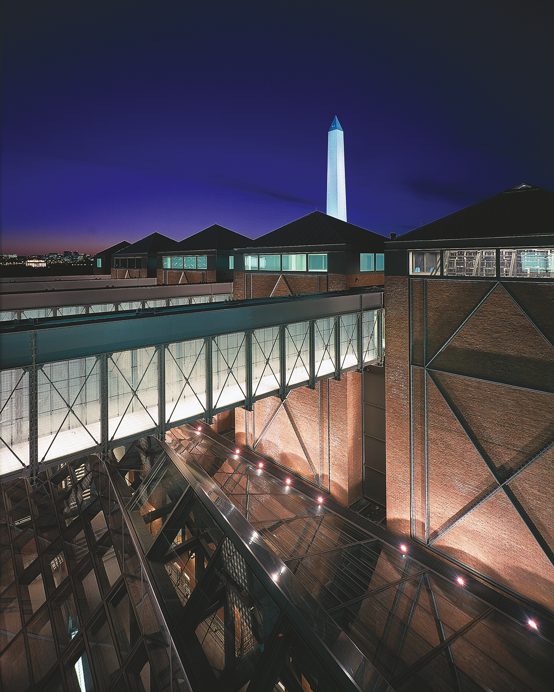 Photograph of brick building exterior with skywalks over a smaller building below with a metal and glass roof and a tall monolith in the background.
