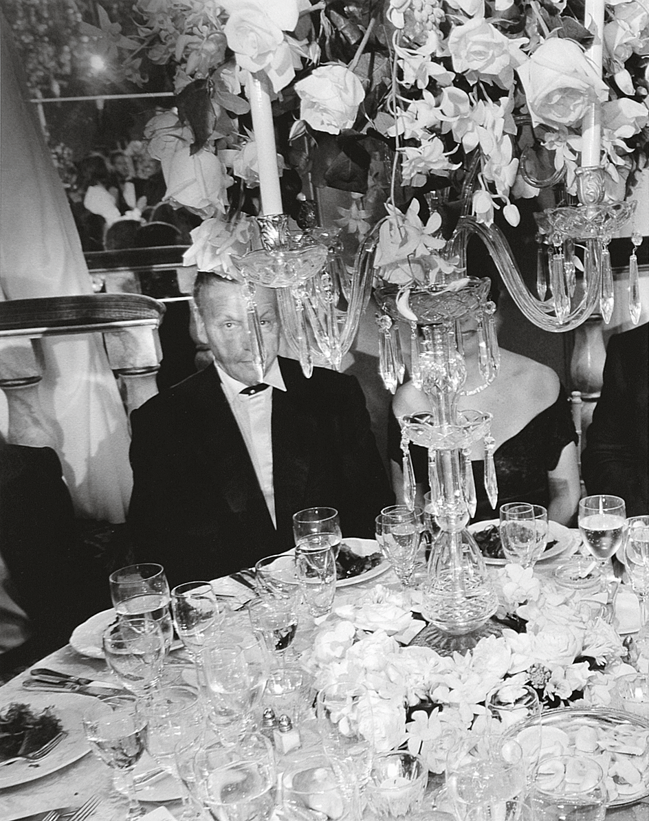 Photograph featuring man seated at a dinner table covered with dishes with his face partially covered by a chandelier.