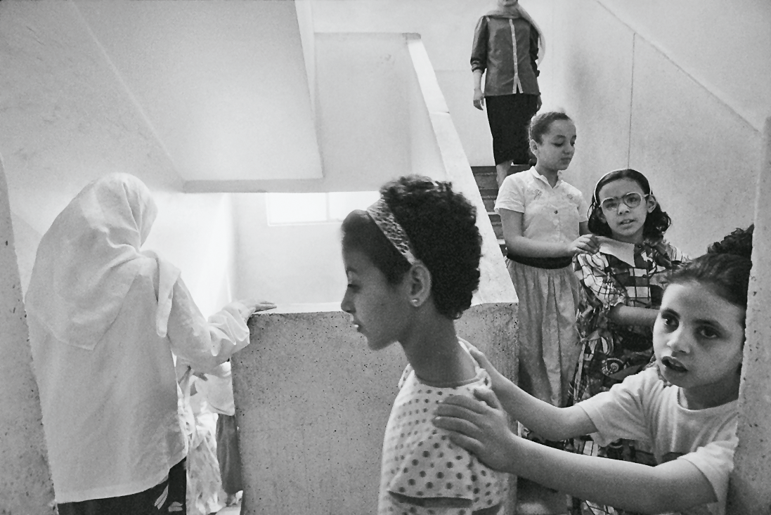 Photograph featuring four young girls holding onto each other's shoulders walking down the stairs in a train with two older women in the front and back.