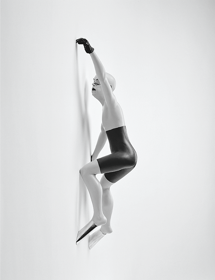Painted bronze figurine climbing wall with no hair or shirt and silver-covered feet; tattoo of figure climbing.