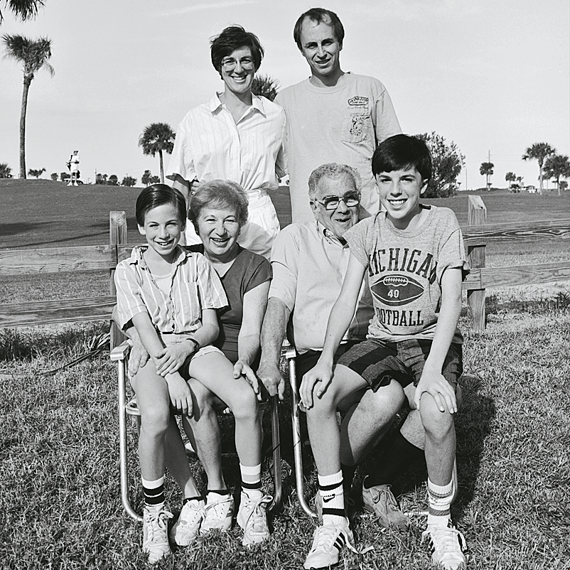 Photograph of two elderly people sitting with two kids in their laps, and two adults standing behind them, all looking at viewer and smiling, with grass and palm trees in background.