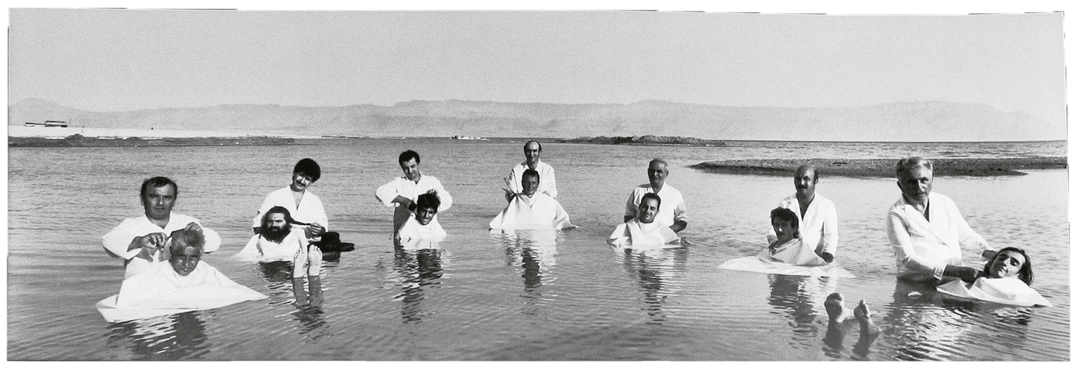 Photograph featuring seven barbers standing in the ocean and cutting hair of seven men floating in front of them. 