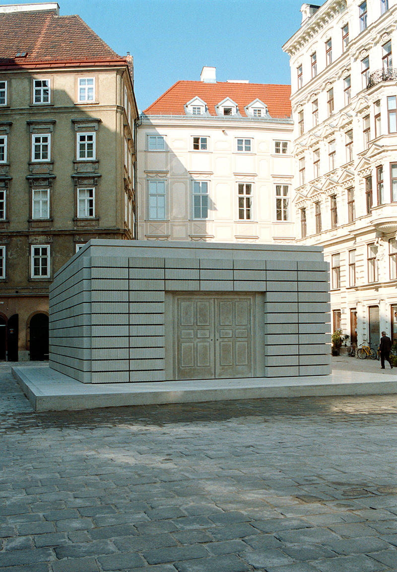 Small rectangular building made of steel and concrete with double doors on square base next to city buildings.