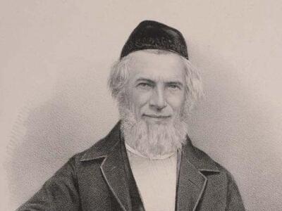 A man with a white beard, a black kippah, and a black frock coat is depicted sitting with his right elbow on a table.