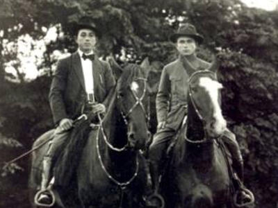 Two men wearing jackets and hate are seated on horses.
