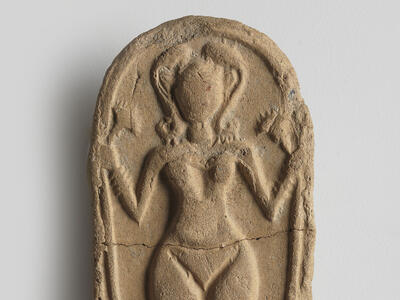 Oval clay piece with figurine of naked woman decorated with headdress, necklace, bracelets, and anklets, and holding a lotus flower.