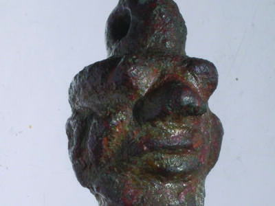Bronze amulet of head with human and animal features and loop on top.