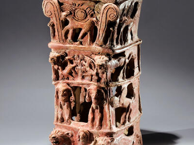 Ceramic offering stand with flat top and four layers of decoration: bottom layer of nude female with raised arms flanked by lions, second layer of two winged figures, third layer of goats eating leaves off tree, and fourth layer of four-legged animal with winged sun disk and palm fronds. 