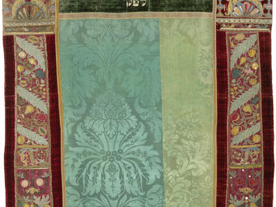 Embroidered curtain of columns with crowns on left and right sides and decorative floral motifs throughout, and Hebrew text in top center.