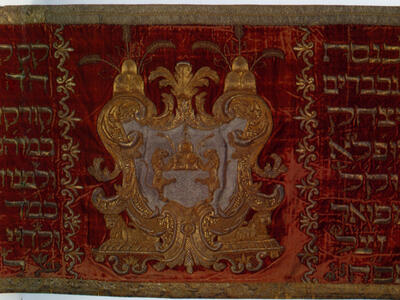 Cloth with embroidered crest in the center with Hebrew text on either side, with floral motifs on left and right edges. 