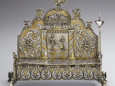 Silver oil lamp with row of eight cups on bottom, and elaborate carvings of two figures and floral designs on lamp with carved birds on top. 