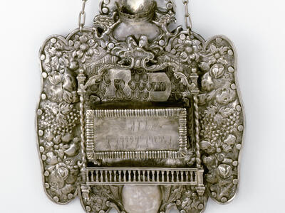 Engraved shield of flowers, grapes, and other decorative elements around rectangular Hebrew inscription.