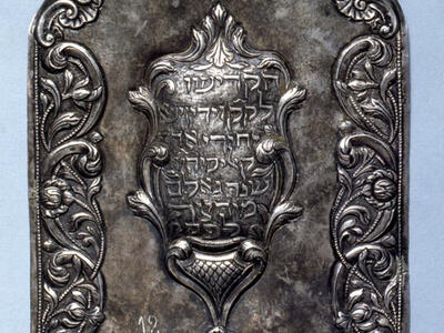 Silver shield of decorative border with Hebrew text in center.