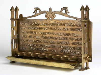 Oil lamp with row of eight cups at the bottom carved with Hebrew inscription and animal heads, and sides resembling buildings.