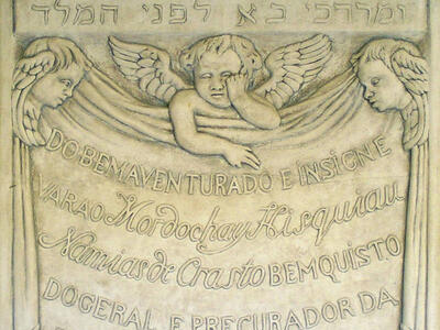 Tombstone of three cherubs and central Portuguese inscription, with engraving of wilted flower, skull, hourglass, and sun, with top panel of engraving of man riding in a procession through street and Hebrew text across the middle. 