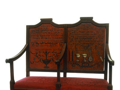 Two-seater bench with central armrest, decorated on the seat backs with ritual objects and Hebrew text.