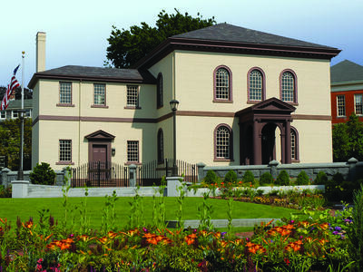 Photograph of exterior of building with semi-circular windows featuring American flag on left and flower garden in front.