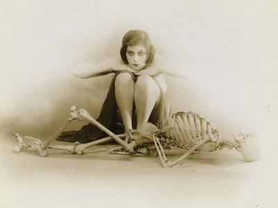 Photograph of female figure sitting behind skeleton.