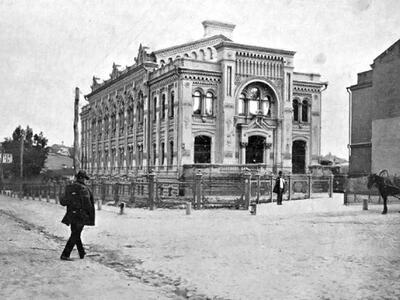 Photograph of exterior of building with rounded window frames and decorative columns, and man in hat walking toward it.