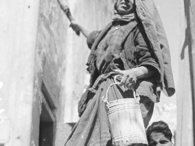 Photograph of woman in headscarf looking toward camera, holding bucket and paintbrush next to wall, with woman behind her looking at camera.