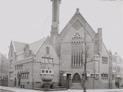 Photograph of a building with triangular roof sections, many long windows, and tower on the corner of a street.