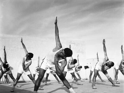 Photograph of rows of men in athletic clothing bending at the waist with their right arms pointed up and their left arms bent in an angle toward their face.