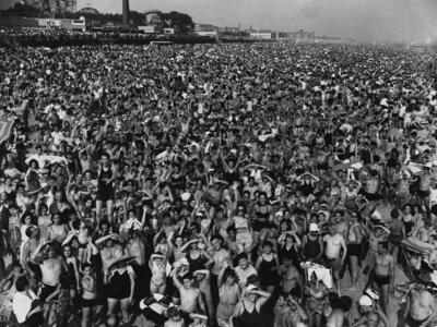 Aerial photograph of hundreds of people crowded together on beach with amusement park in background.