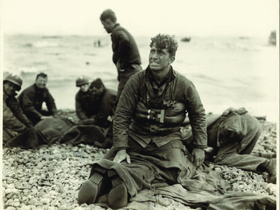 Photograph of soldiers on the beach, with man in center facing the viewer and looking up, as he kneels on top of a prone soldier.