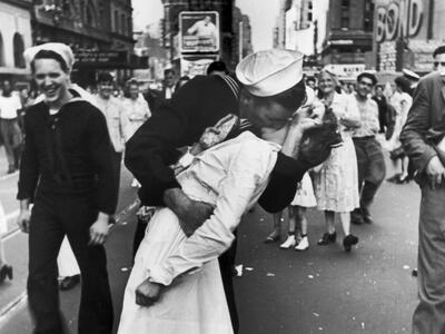 Photograph of sailor grabbing and kissing a woman in a busy city street with people and buildings in background. 