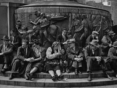 Photograph of men, women, and children seated on the steps of a large public monument with nude classical figures and a winged horse atop a circular base.