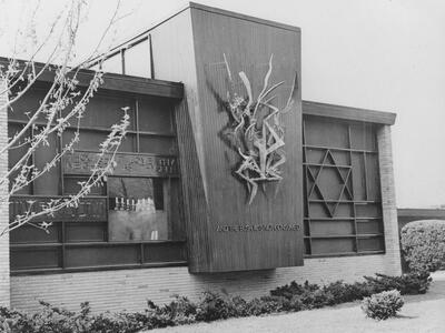 Exterior wall of synagogue featuring Star of David on the right side of the wall and a ribbon-like sculpture in the middle of the wall on a prominent vertical section that juts out from the rest of the wall, with inscription in English reading "And the bush was not consumed."