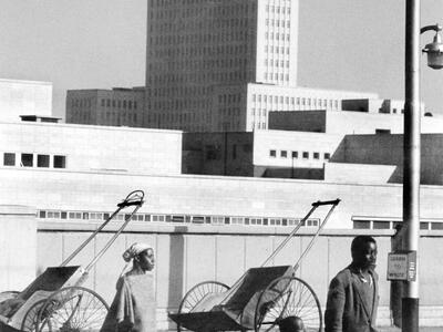 Photograph of man, woman, and child walking down city street with high-rise building and two rickshaws in background.