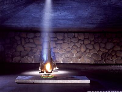 Memorial depicting flame burning from a base and illuminating a wall with an angular roof and basalt walls, with place names in English and Hebrew on the floor.