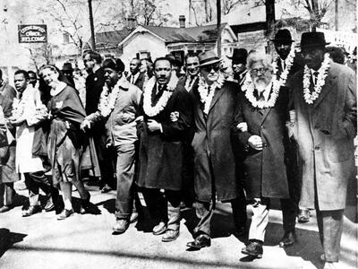 Photograph of men marching down the street with arms linked wearing white double carnation leis. 