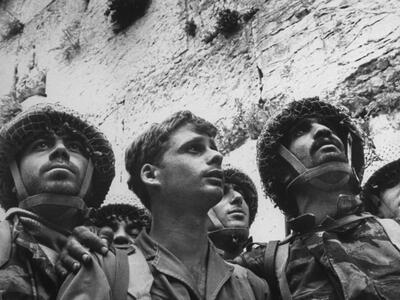 Photograph of three soldiers in uniform looking to their left standing in front of the Western Wall, with central soldier holding helmet in his hands.
