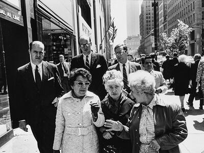 Photograph of three elderly women walking and talking on busy city sidewalk with men in suits behind them.