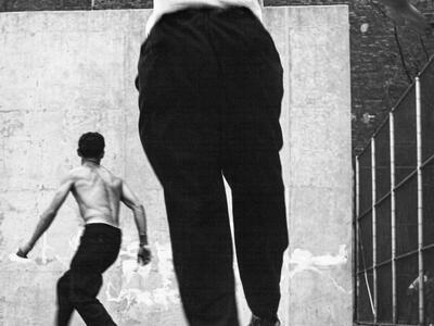 Photograph of two men playing handball, both of whom are turned away from the viewer and facing the wall. 