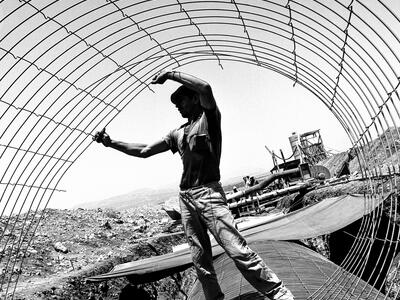 Laying the Pipes of the National Water Carrier from the Sea of Galilee to the Negev Photograph of man holding a wire cylinder and standing on a large pipe, with a construction site behind him.
