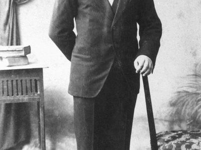 Photograph of man in suit with one arm on a walking stick. 