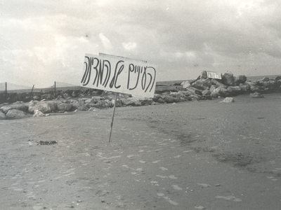Photograph of Hebrew sign on empty beach with a rock jetty in background.