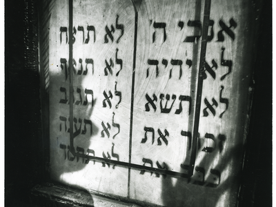 Photograph featuring shadow of a face and a hand over a wall with Hebrew text. 