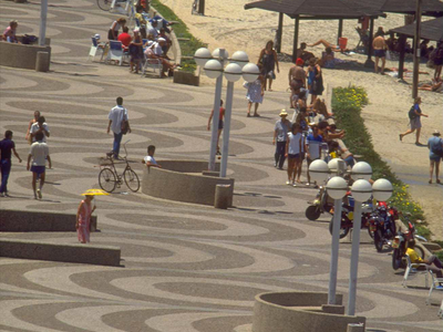 Photograph featuring a promenade alongside a beach with people walking and sitting, with several sets of lightposts and circular benches.