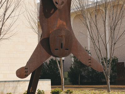 Steel sculpture on steel frame outside of building. Top of sculpture is the head of a sheep with horns and bottom of sculpture is stylized head and arms of a human figure.