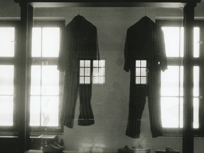 Photograph depicting two jumpsuits hanging between two large windows superimposed over an image of a human figure standing with their back turned. 